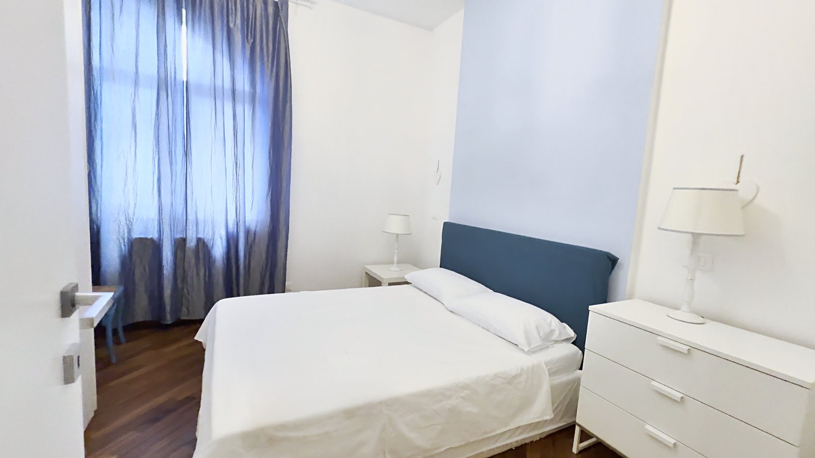 Schlafzimmer mit Doppelbett, blauem Kopfteil, weißer Kommode und blauen Vorhängen, Via Machiavelli, Triest