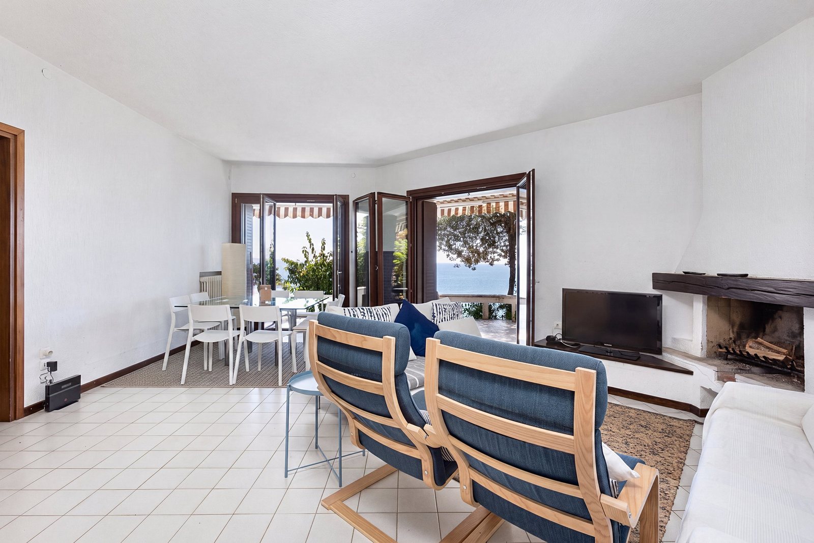 Living room with fireplace, two blue armchairs, dining table and French windows onto the sea-view terrace