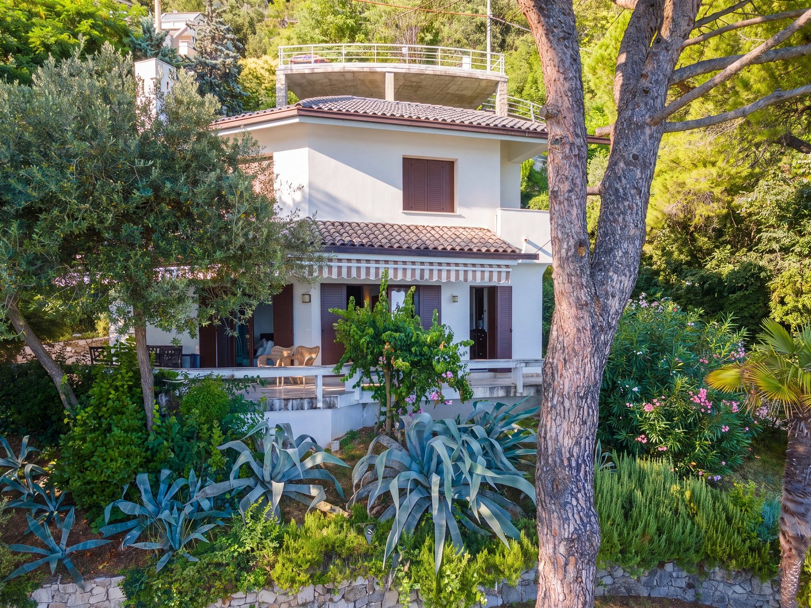 Villa facade with terrace and striped awning, agaves and pines in the garden
