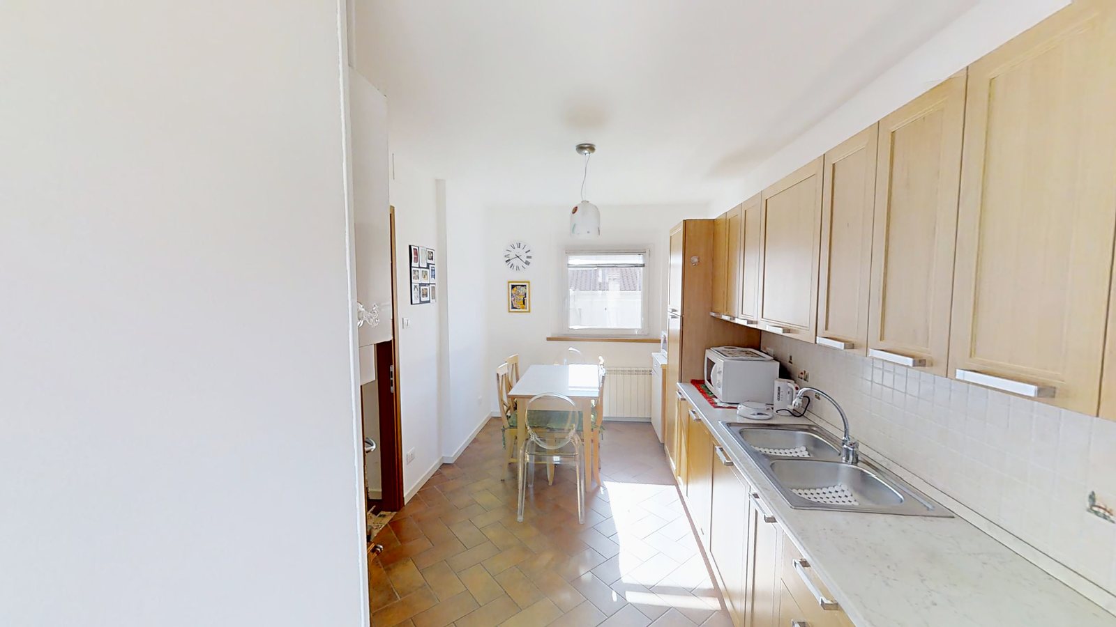 Küche mit Tisch vor dem Fenster, Wanduhr und Hängeschränken aus hellem Holz, Via Slataper, Triest