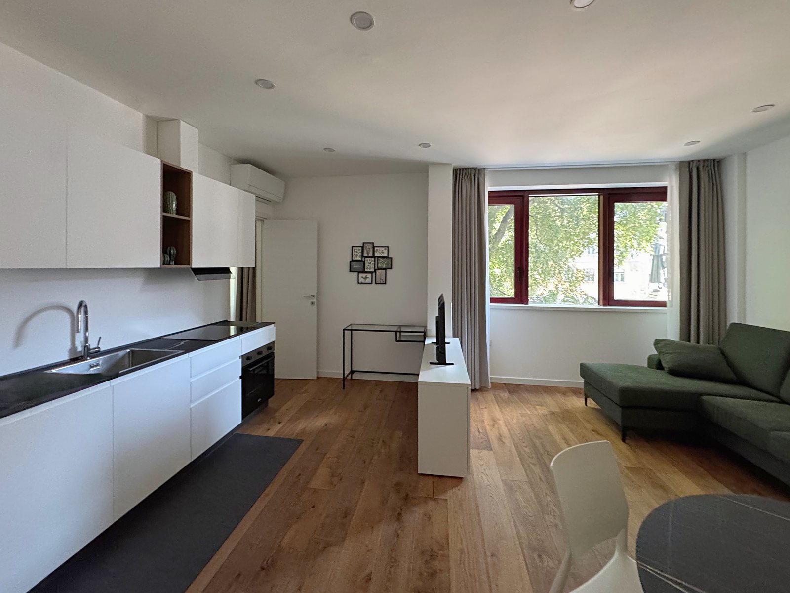 Wohnbereich mit weißer Küche, grünem Sofa und Fenstern zu den Bäumen, in der Wohnung in der Via Marconi, Triest