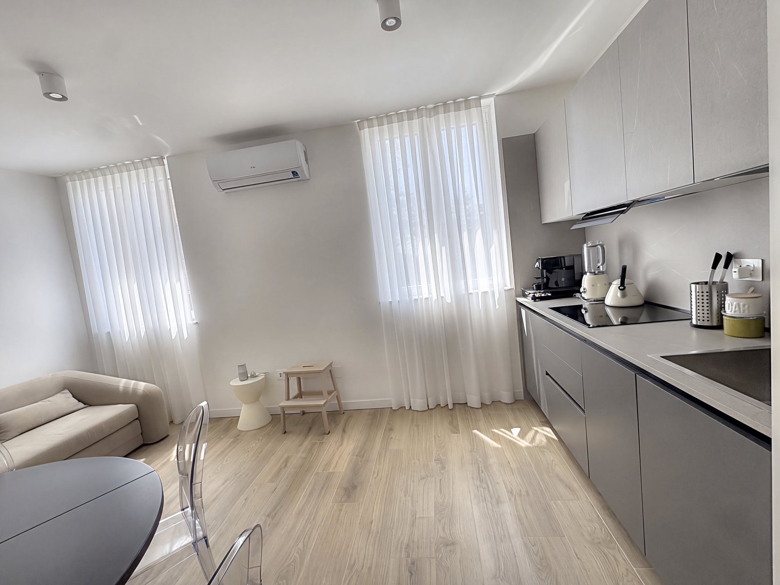 Living area with grey fitted kitchen, sofa and two windows with white curtains, Salita di Gretta, Trieste