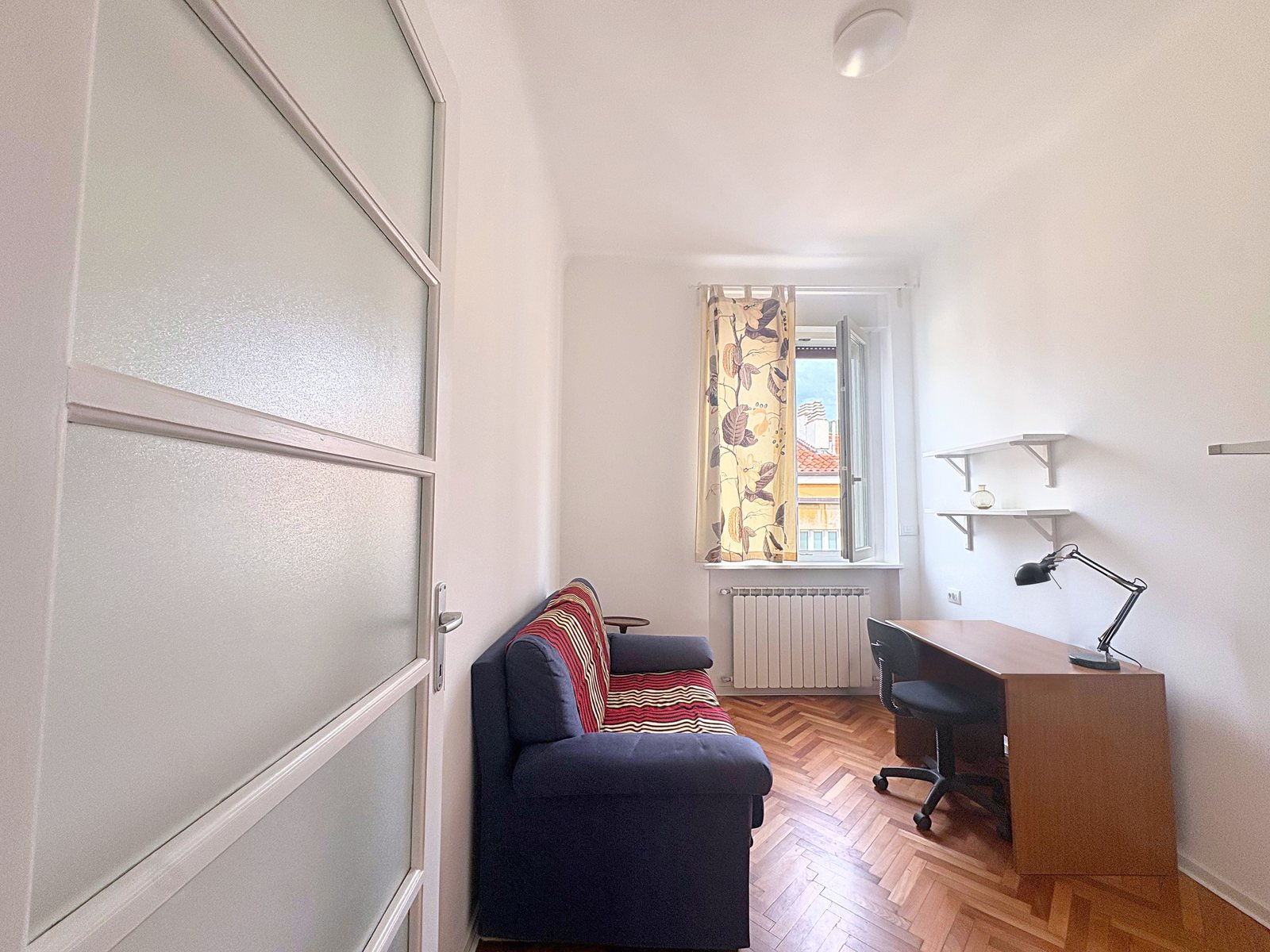 Stanza piccola con divano, scrivania in legno, mensole bianche e finestra con tenda a fiori, Trieste