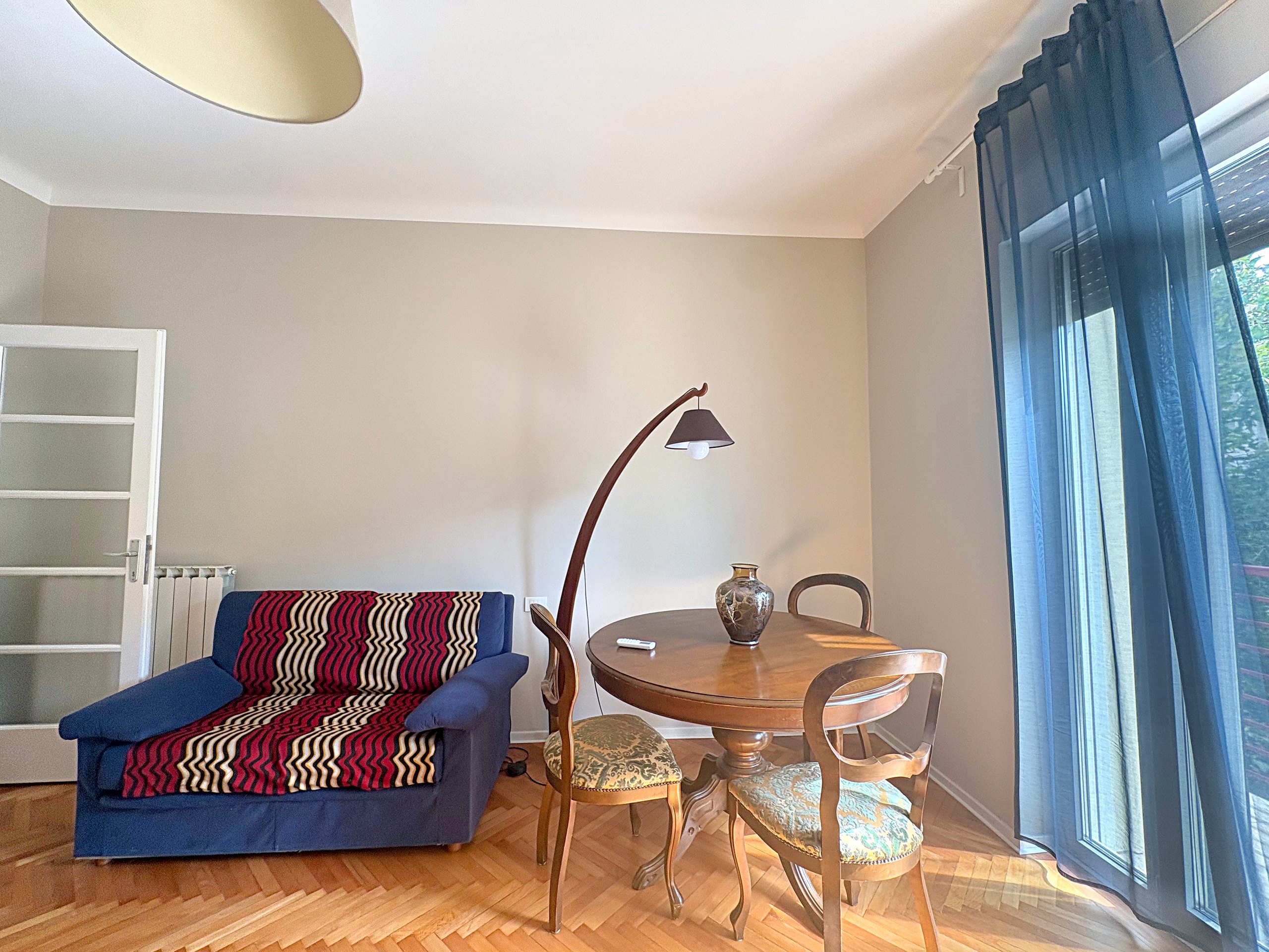 Stanza grande con divano blu, tavolo rotondo in legno e lampada ad arco accanto alla porta-finestra, Trieste