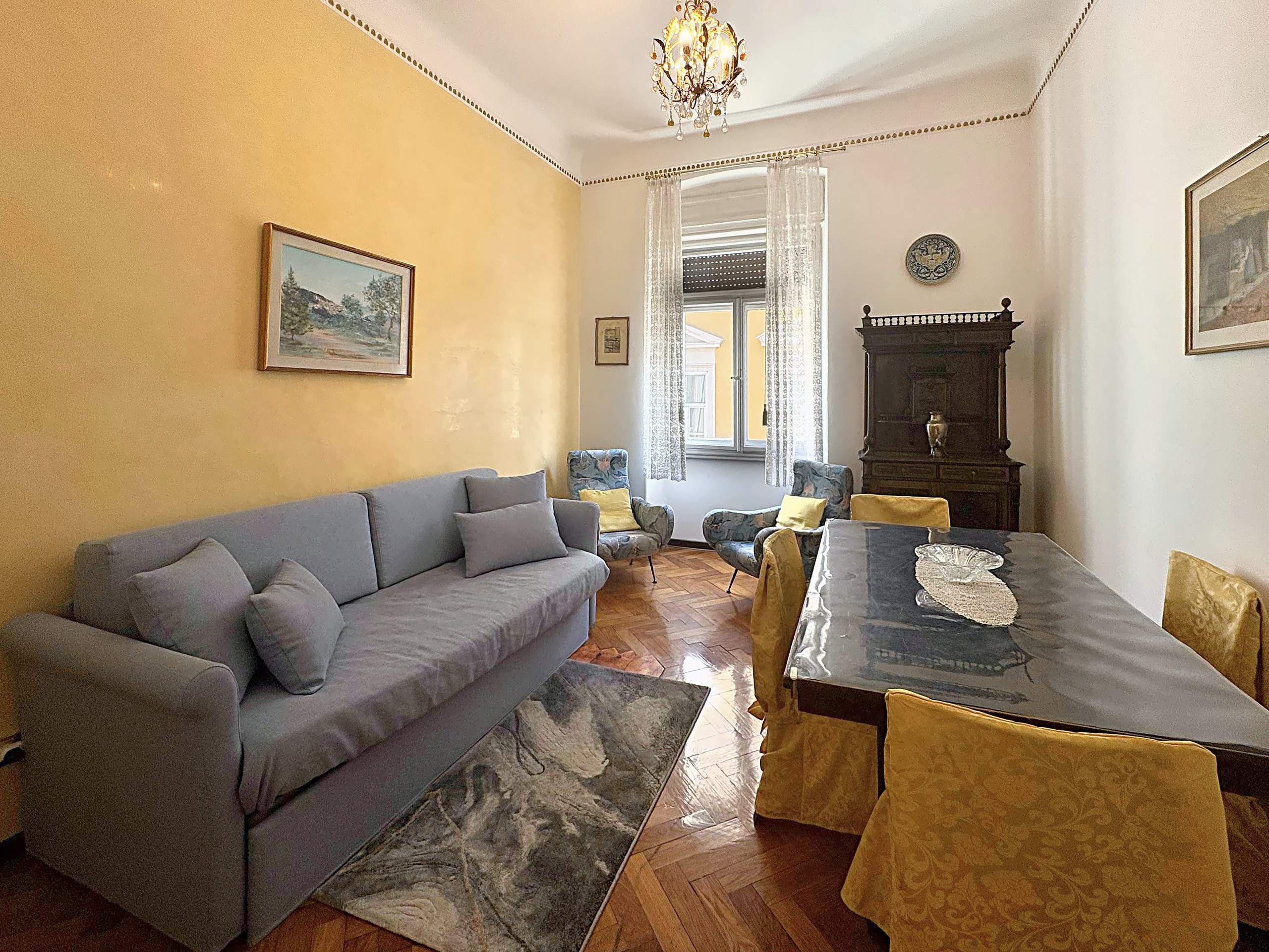 Soggiorno con parete gialla, divano grigio, tavolo da pranzo e lampadario di cristallo, via San Giorgio, Trieste