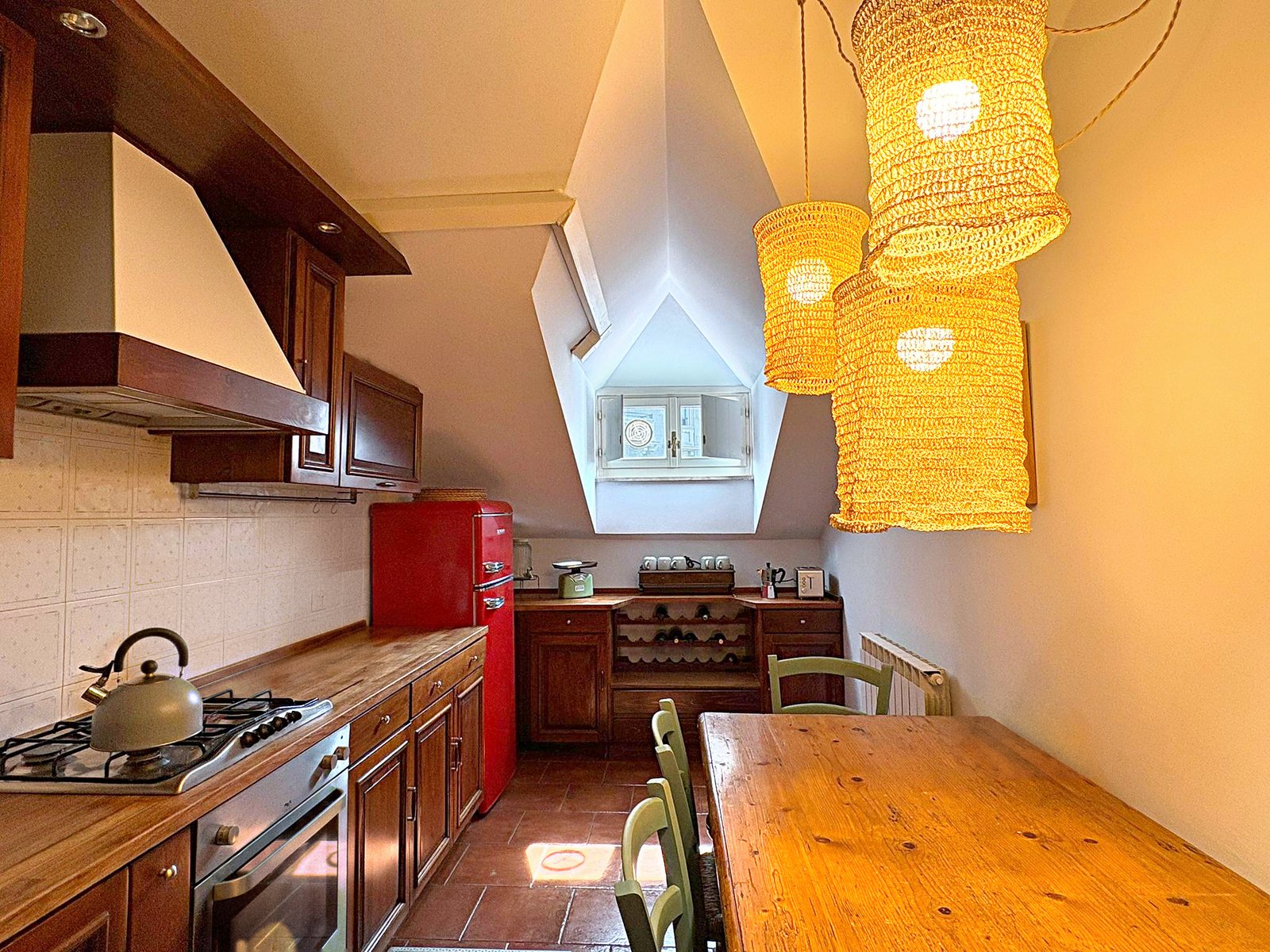 Cucina in legno con frigorifero rosso, lampade in vimini e tavolo sotto l'abbaino, mansarda a Trieste
